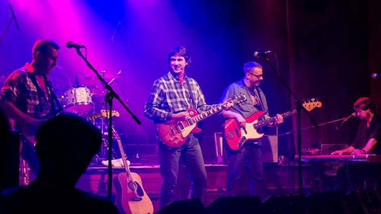 Die Band auf der Bühne in blau-violettem Licht, vorne zwei Gitarristen