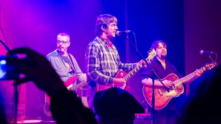 Drei Musiker mit Gitarren in violettem Licht, vorne eine Hand mit Handy aus dem Publikum